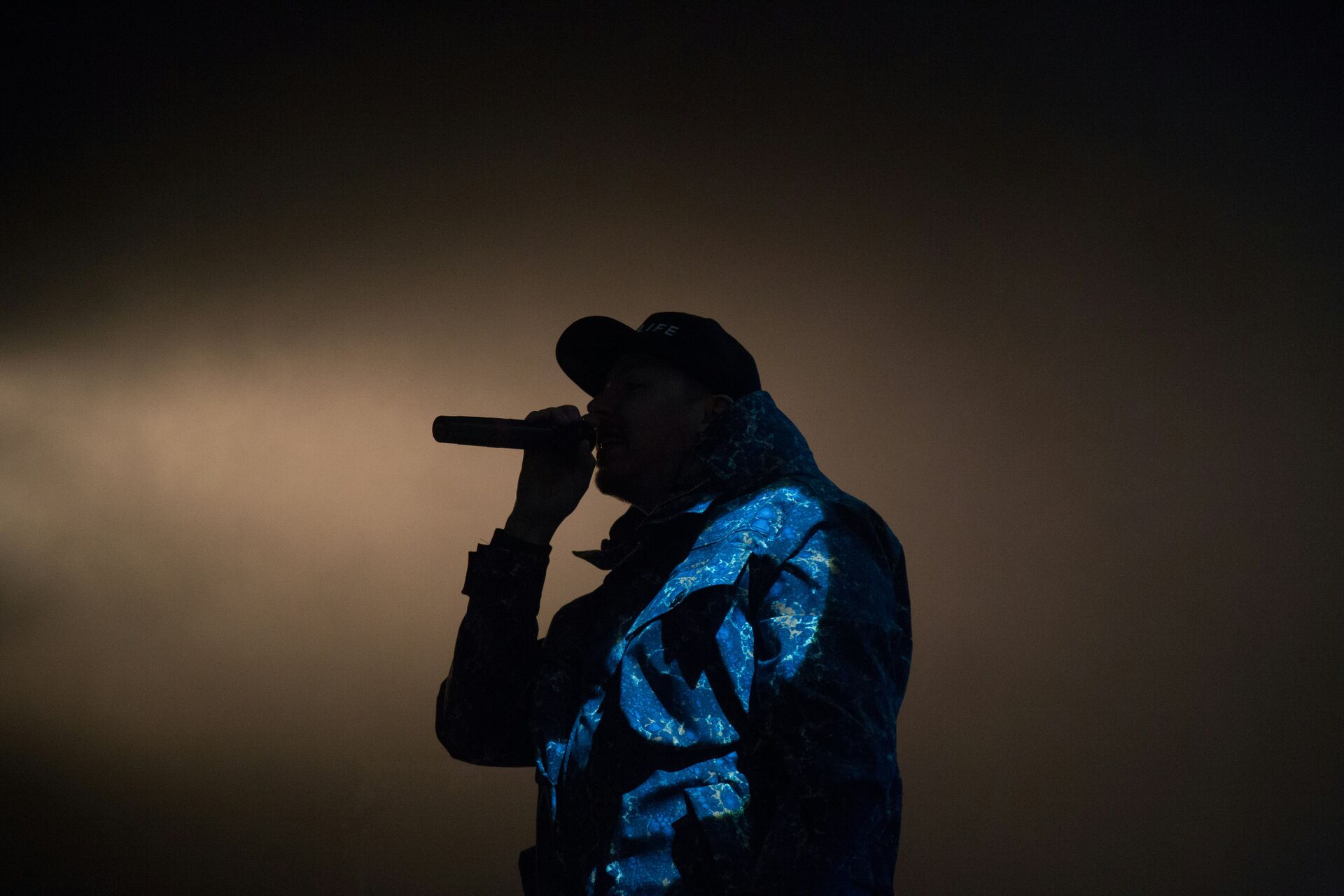Performer in a blue patterned jacket singing into a microphone on a dimly lit stage.
